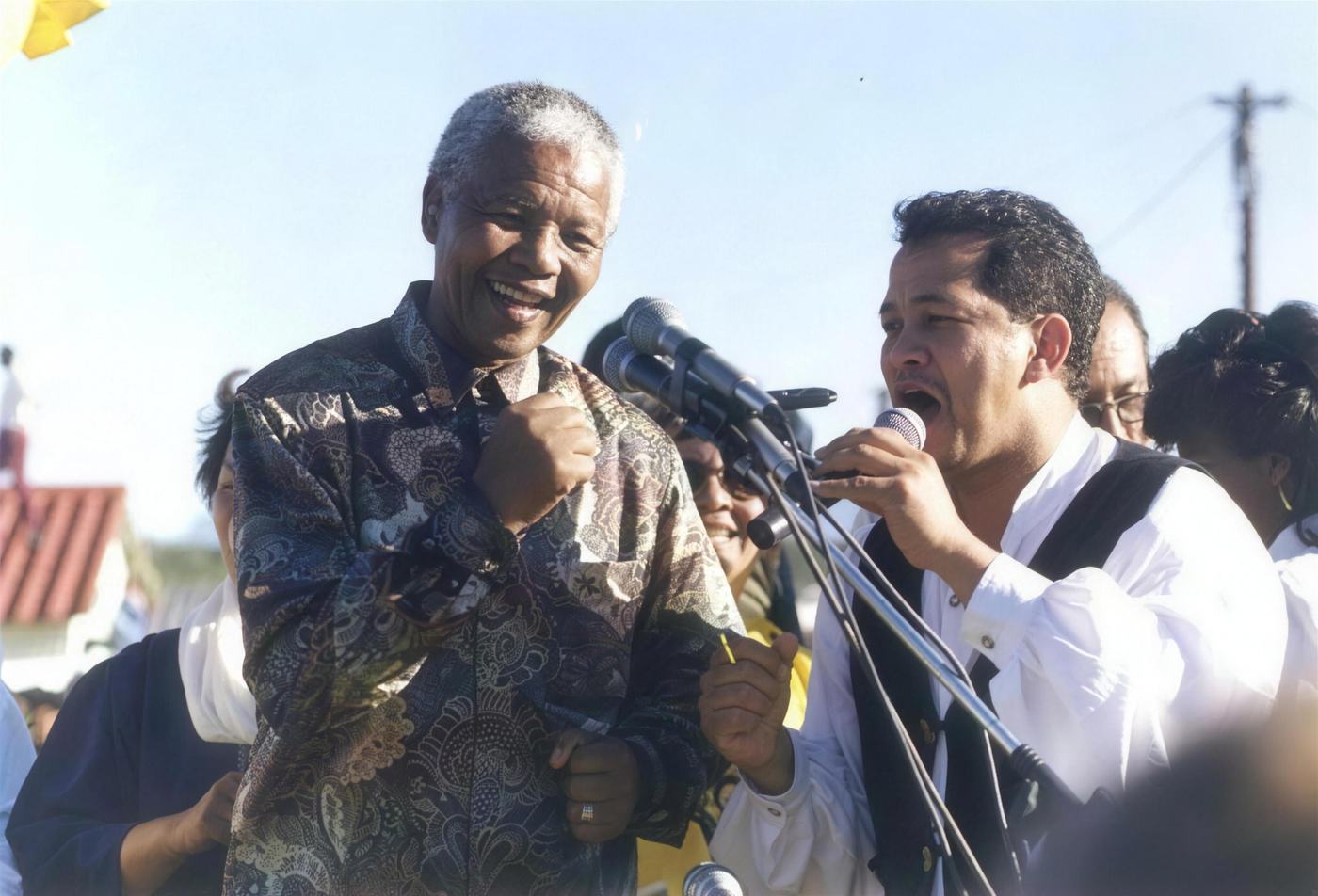 Nelson Mandela laughing beside John Pretorius singing at a bank of microphones