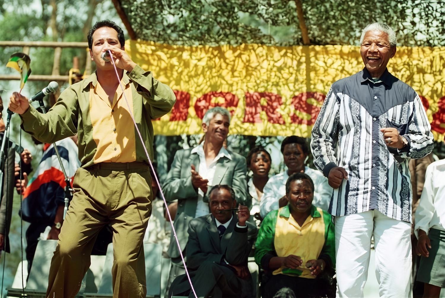 John Pretorius singing as Nelson Mandela moves to the music beside him, under a yellow banner
